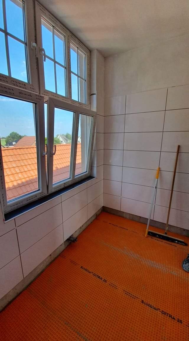 Wnętrze w trakcie prac wykończeniowych: białe płytki na ścianach, pomarańczowa mata drenażowa Schluter-DITRA 25 na podłodze, widok przez okno na dachy domów.