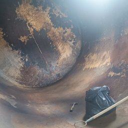 KOLORTEK - TOMASZ TWARDOSZ - Wnętrze metalowego zbiornika po piaskowaniu, z resztkami starej farby i narzędziami: młotek, grabie i worek na śmieci. Widok z dołu, ukazujący skalę i zakres prac.