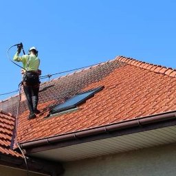 KOLORTEK - TOMASZ TWARDOSZ - Pracownik w uprzęży myje dach z czerwonej dachówki za pomocą myjki ciśnieniowej. Część dachu jest już czysta, widoczne okno dachowe i brązowa rynna.