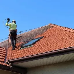 Pracownik w uprzęży myje dach z czerwonej dachówki za pomocą myjki ciśnieniowej. Część dachu jest już czysta, widoczne okno dachowe i brązowa rynna.