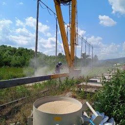 KOLORTEK - TOMASZ TWARDOSZ - Pracownik w kasku piaskuje metalową konstrukcję przy użyciu specjalistycznego sprzętu. W tle widoczne drzewa, niebo i elementy konstrukcyjne.