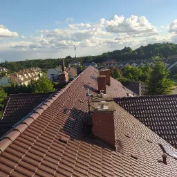 Czerwono-brązowy dach z dachówki ceramicznej z kominami na tle zabudowy jednorodzinnej i błękitnego nieba.