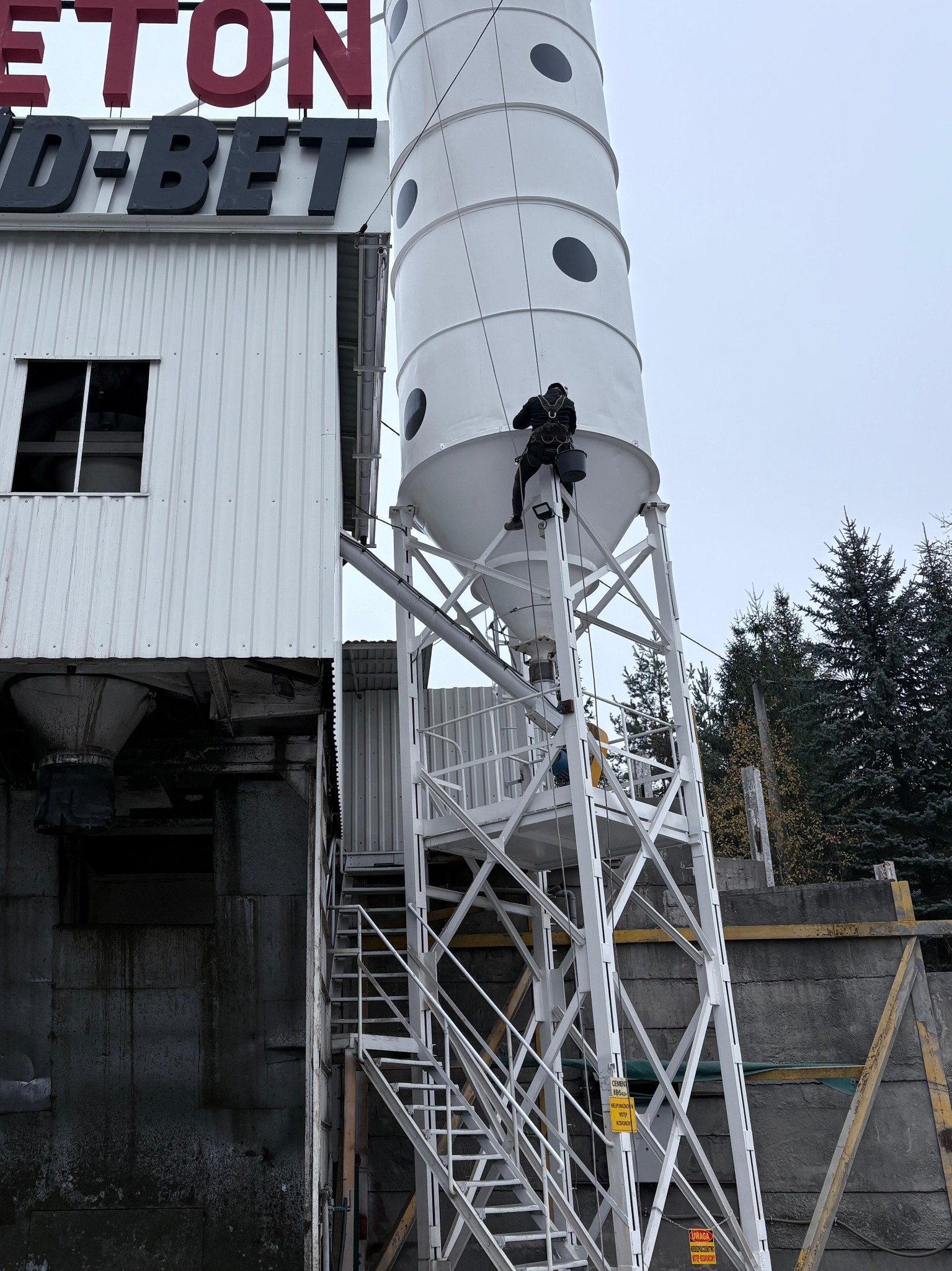Pracownik na linie zabezpieczającej czyści silos na terenie zakładu produkcyjnego betonu. Widoczne elementy konstrukcji stalowej i drabiny.