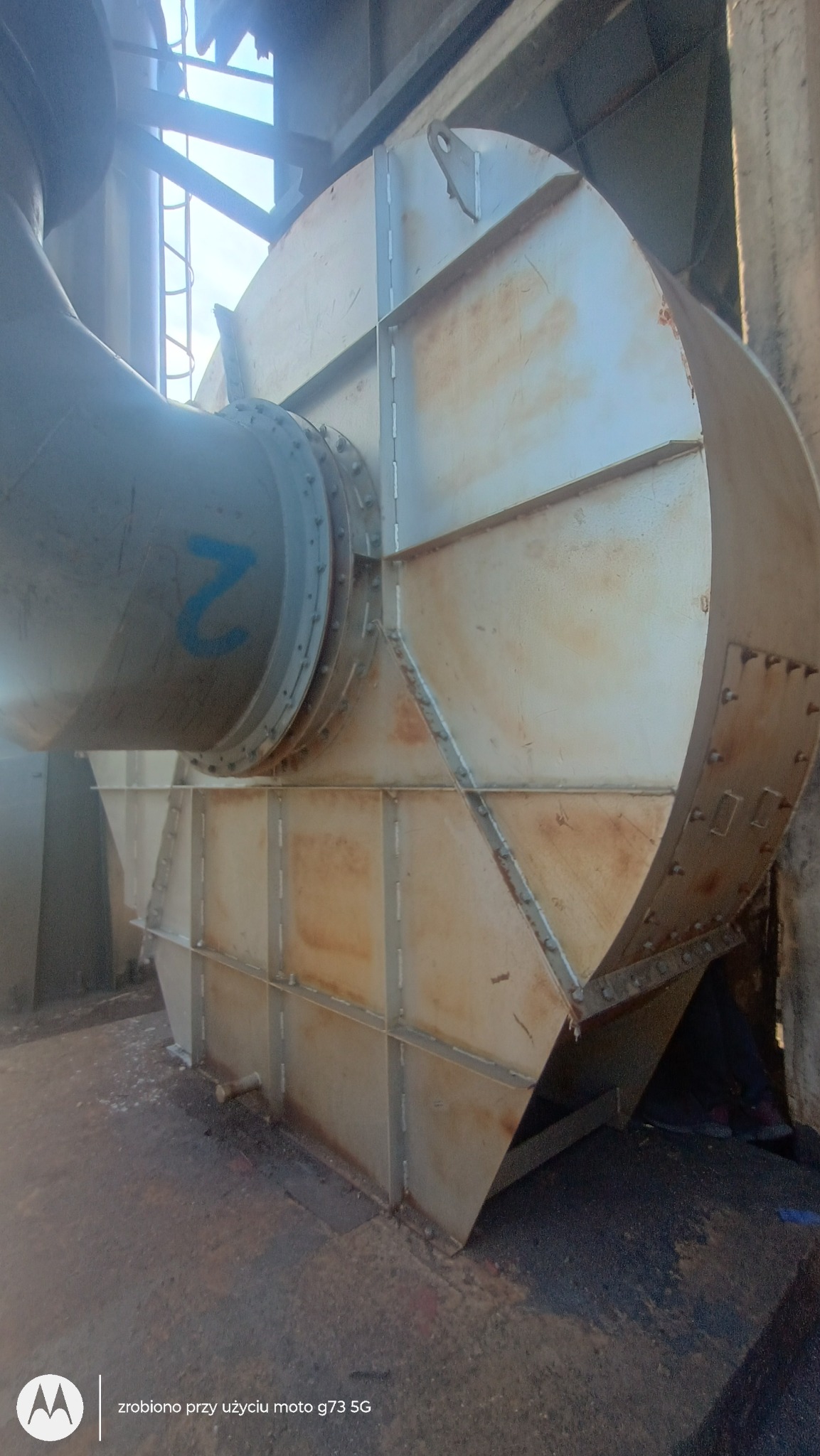 Metalowa konstrukcja wentylacyjna z rurą, widoczne ślady korozji i spawania. Ujęcie z dołu, podkreślające wielkość i złożoność instalacji.