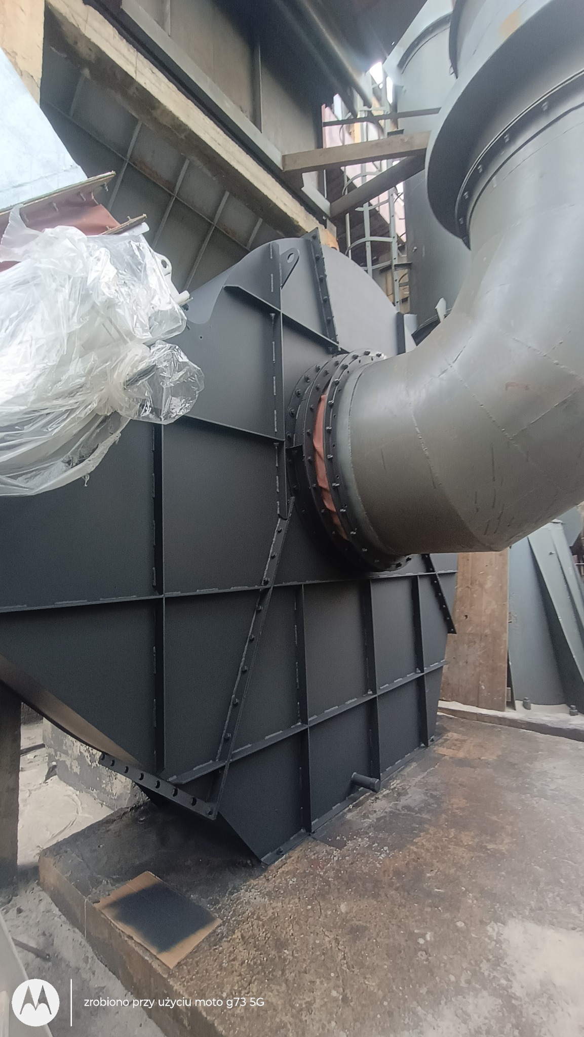 Konstrukcja przemysłowa po malowaniu: czarna, metalowa obudowa z rurą w kolorze szarym. Widoczne ślady farby na podłożu. Ujęcie z dołu.