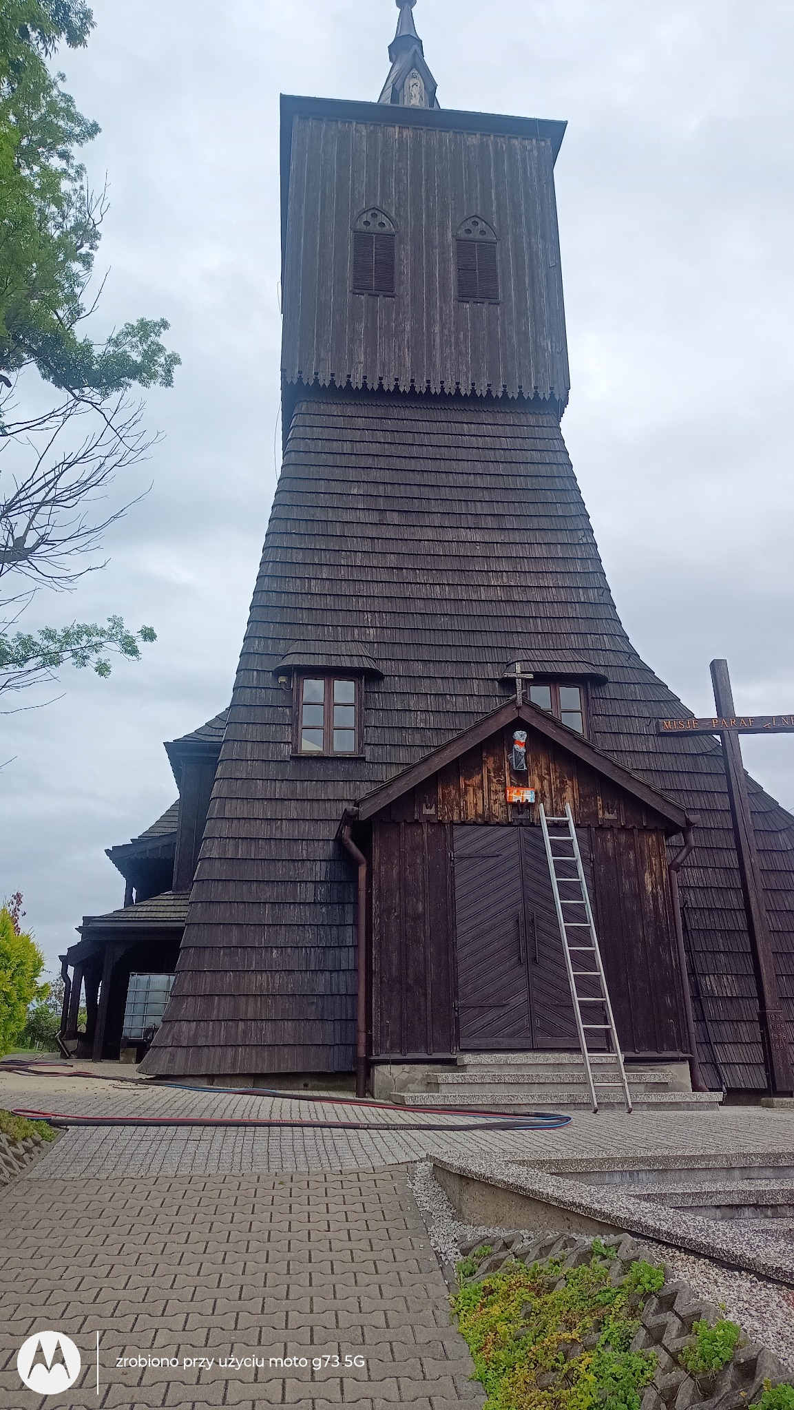 Drewniany kościół kryty gontem z drabiną opartą o wejście. Wokół brukowany chodnik i zieleń. Widoczne węże do mycia.