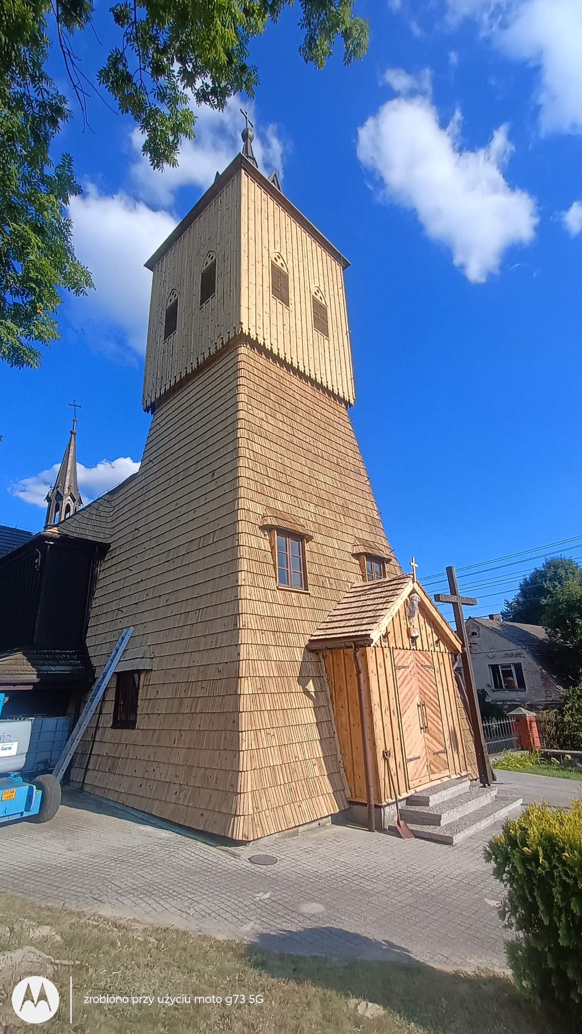 Drewniany kościół z nową elewacją z gontów, wieża z ażurową konstrukcją, błękitne niebo. Widoczny fragment dachu sąsiedniej budowli i podnośnik koszowy.
