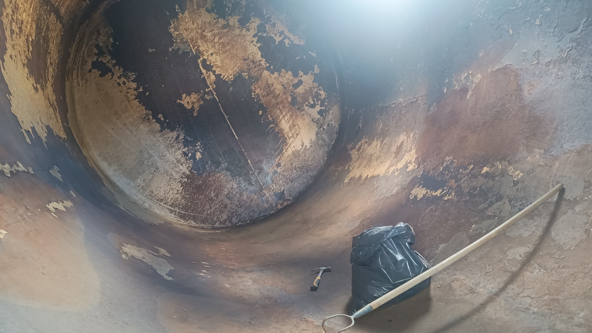 Wnętrze metalowego zbiornika po piaskowaniu, z resztkami starej farby i narzędziami: młotek, grabie i worek na śmieci. Widok z dołu, ukazujący skalę i zakres prac.