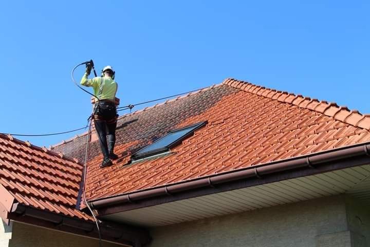 Pracownik w uprzęży myje dach z czerwonej dachówki za pomocą myjki ciśnieniowej. Część dachu jest już czysta, widoczne okno dachowe i brązowa rynna.