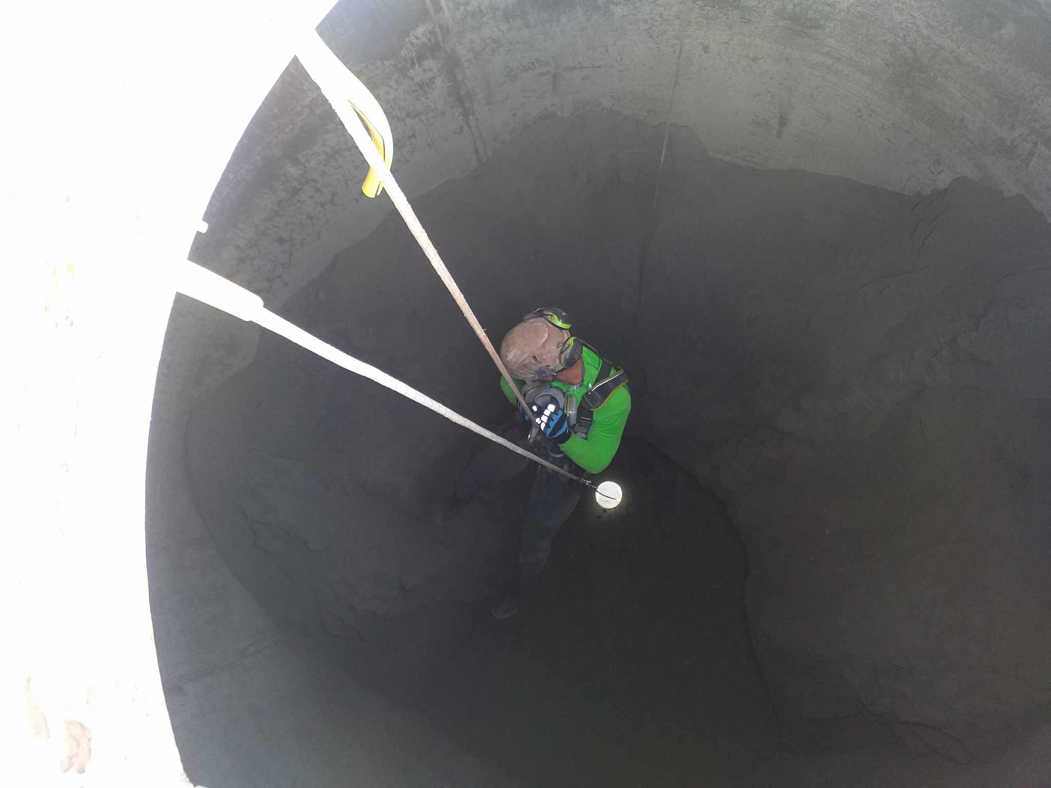 Pracownik w uprzęży, oświetlony, wewnątrz betonowego szybu. Widok z góry ukazuje linę i elementy ochronne. Specyficzne oświetlenie i surowa faktura betonu.