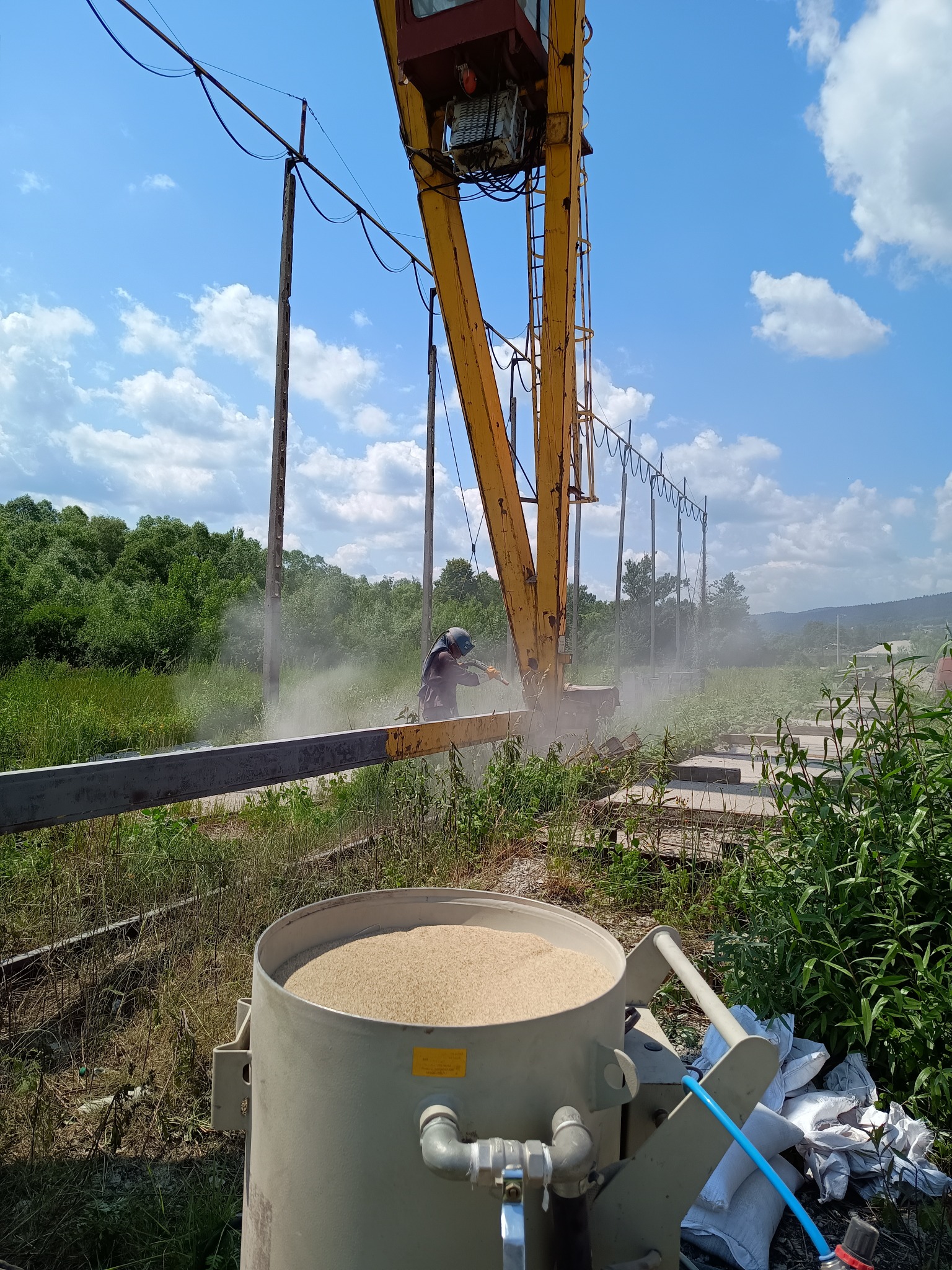Pracownik w kasku piaskuje metalową konstrukcję przy użyciu specjalistycznego sprzętu. W tle widoczne drzewa, niebo i elementy konstrukcyjne.