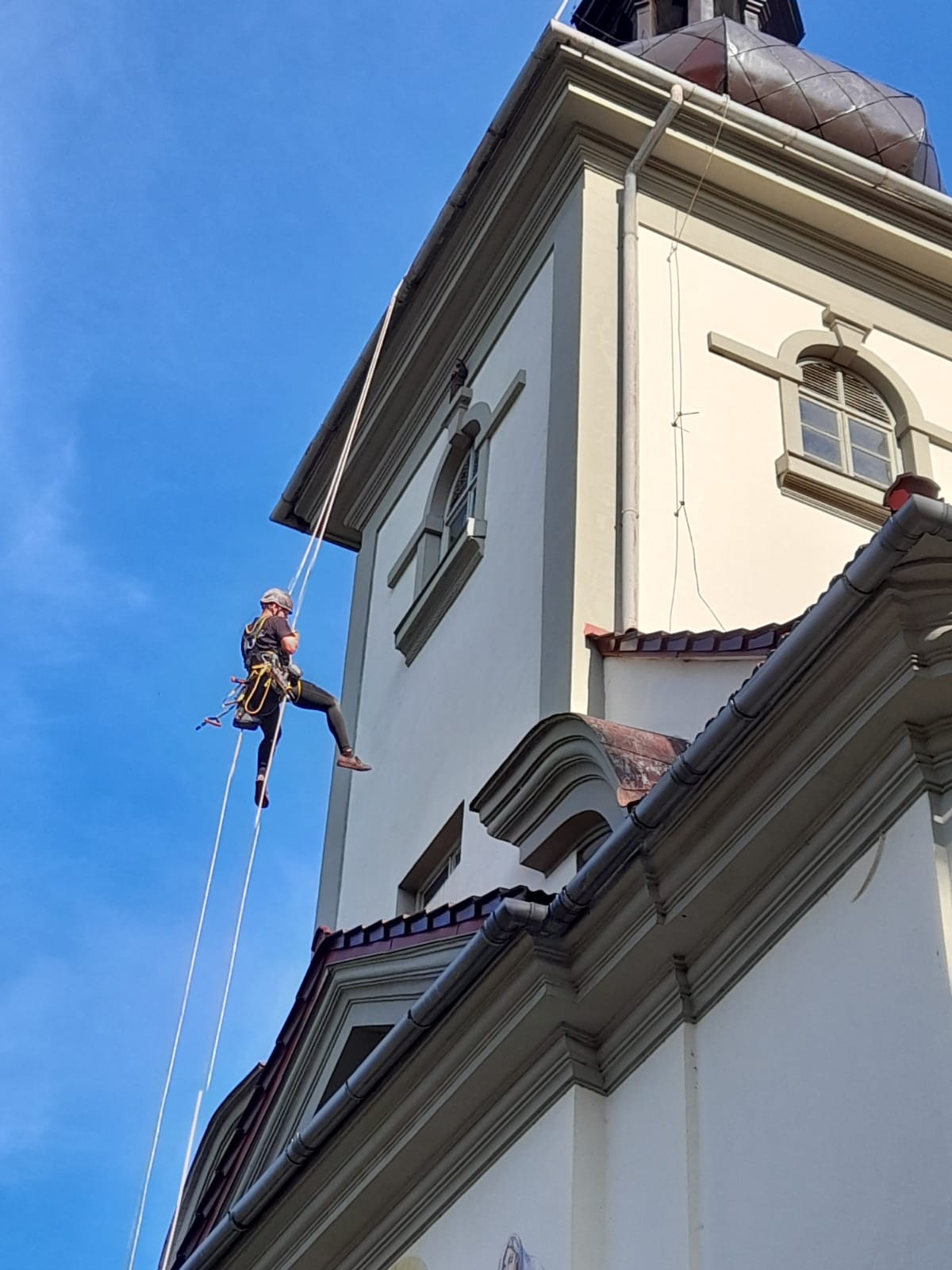 Pracownik na linach zabezpieczających, wykonujący prace na wysokości na budynku z rynnami. Widoczny fragment dachu i elewacji. Błękitne niebo w tle.