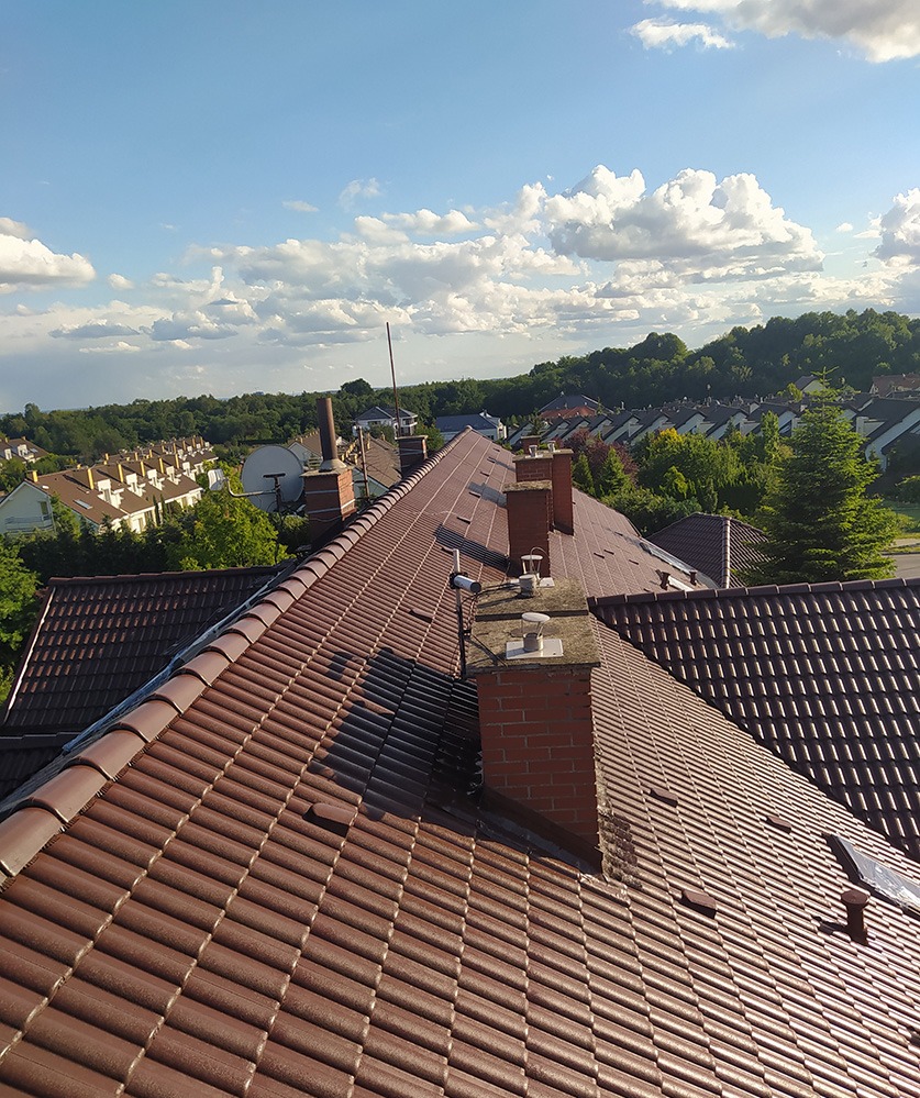 Czerwono-brązowy dach z dachówki ceramicznej z kominami na tle zabudowy jednorodzinnej i błękitnego nieba.