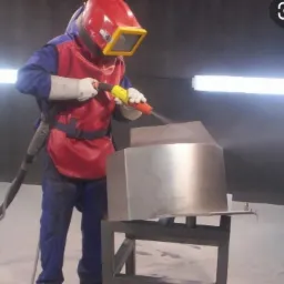 Pracownik w kombinezonie ochronnym i masce piaskuje metalowy element na stojaku w warsztacie, strumień ścierniwa skierowany na obrabianą powierzchnię.