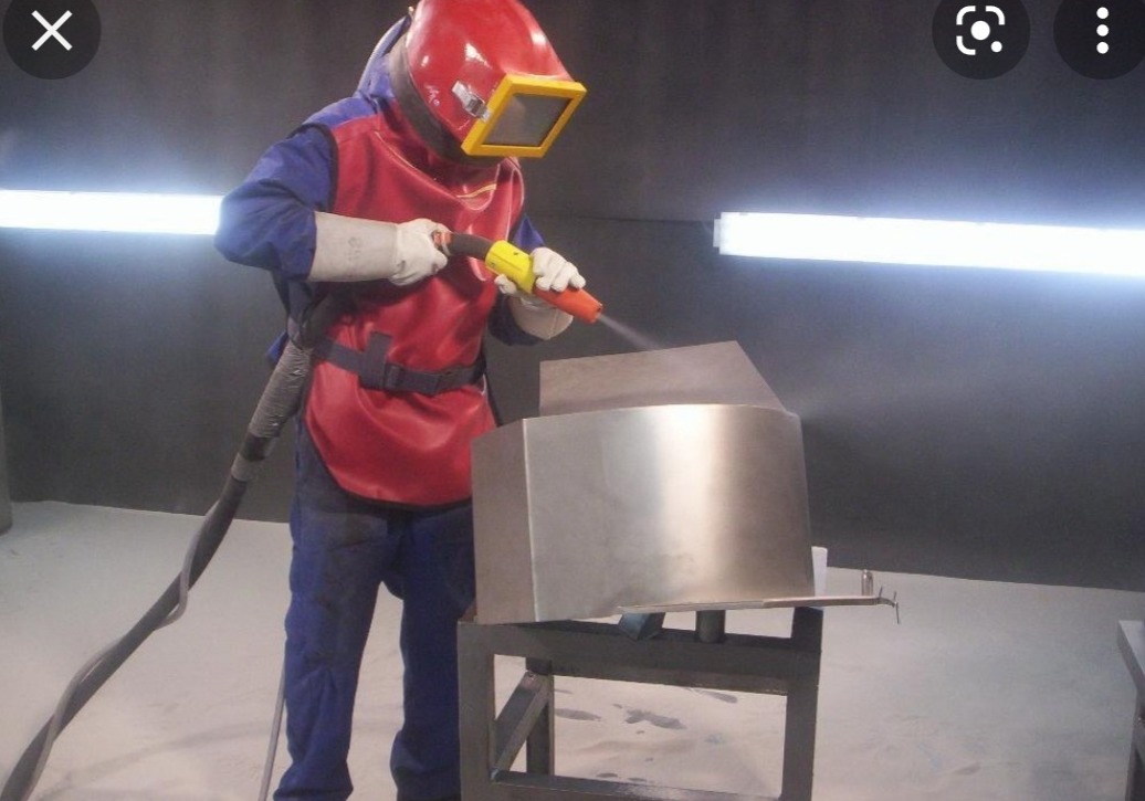 Pracownik w kombinezonie ochronnym i masce piaskuje metalowy element na stojaku w warsztacie, strumień ścierniwa skierowany na obrabianą powierzchnię.