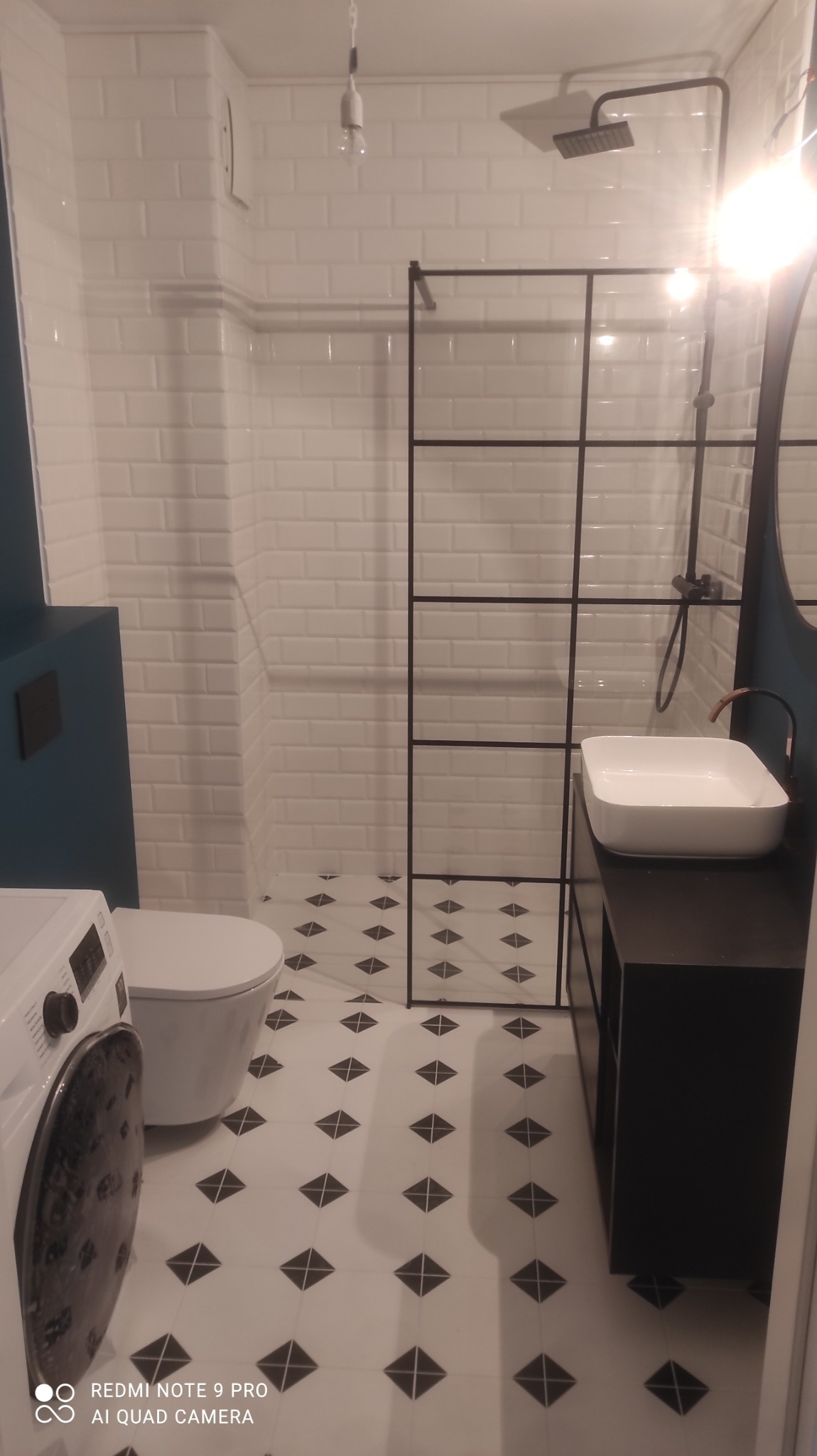 Nowoczesna łazienka z kabiną prysznicową typu walk-in z czarnymi profilami, białą umywalką nablatową na czarnej szafce, białą toaletą i pralką, oraz biało-czarną podłogą w romby.