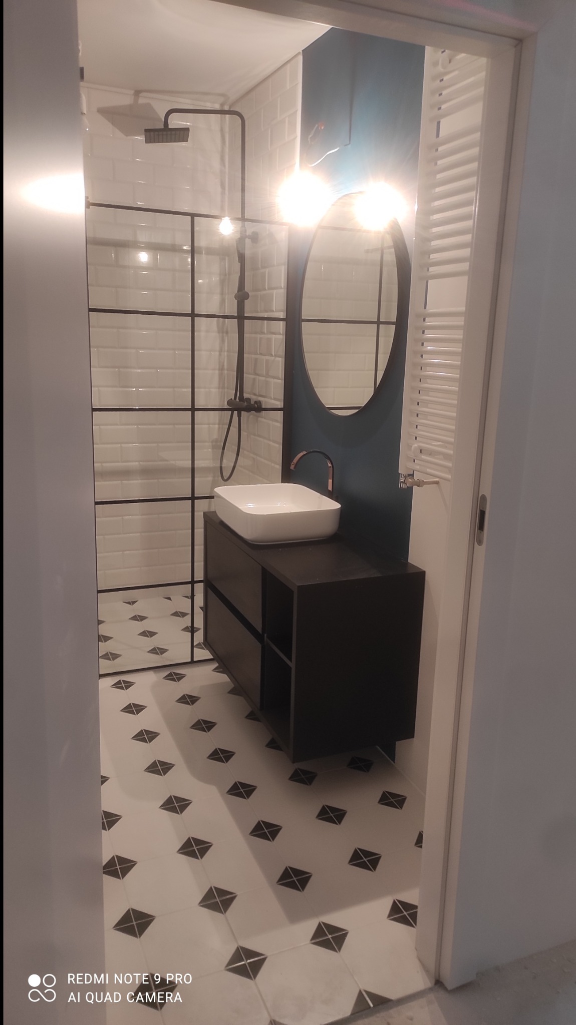 Nowoczesna łazienka z kabiną prysznicową w stylu loft, białą umywalką nablatową na czarnej szafce z szufladami, okrągłym lustrem na granatowej ścianie i grzejnikiem drabinkowym.