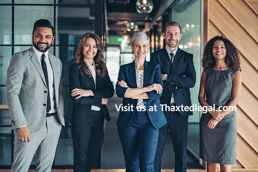 Profesjonalny zespół prawniczy: pięcioro uśmiechniętych prawników w garniturach stojących w nowoczesnym biurze z przeszklonymi ścianami.