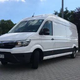 Biały, dostawczy bus marki MAN TGE z polską rejestracją PO 4WR39, zaparkowany na szarej kostce brukowej, z zielenią drzew w tle i błękitnym niebem.