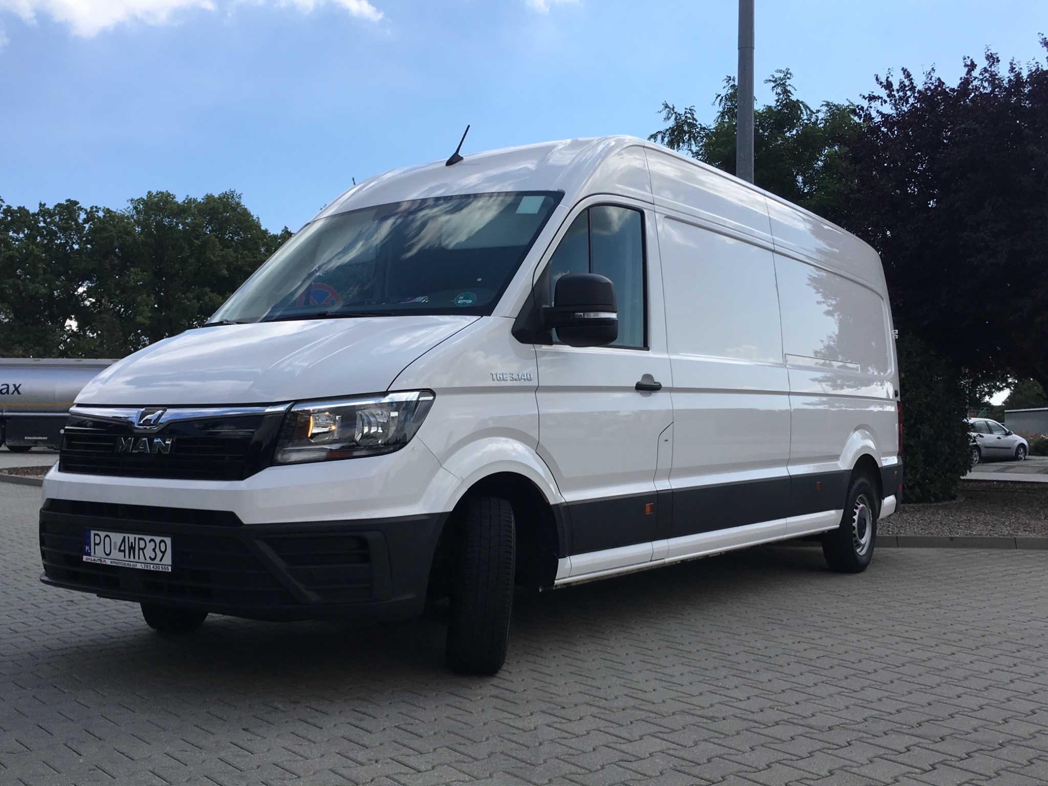 Biały, dostawczy bus marki MAN TGE z polską rejestracją PO 4WR39, zaparkowany na szarej kostce brukowej, z zielenią drzew w tle i błękitnym niebem.