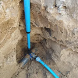 Błękitne rury wodociągowe połączone z ciemną rurą w wykopie ziemnym, widoczne złącza i piasek o różnej wilgotności na ścianach wykopu.