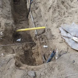Wykop w ziemi z widocznymi rurami: żółtą gazową i czarną kanalizacyjną, oraz łopatą opartą o ścianę wykopu. Widoczne białe sznurki wyznaczające trasę.