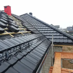 Częściowo pokryty dach czarną dachówką ceramiczną z widocznym rusztowaniem i kominem w czerwonej osłonie, podczas prac dekarskich.