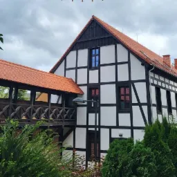 Elewacja domu w stylu szachulcowym z czerwonym dachem i drewnianym łącznikiem, widok z poziomu ogrodu z iglakami.