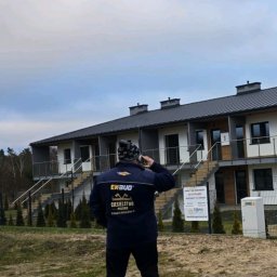 Ciesielstwo Polskie - Mężczyzna w kurtce z logo firmy dekarskiej EKSBUD rozmawia przez telefon na tle szeregu nowo wybudowanych domów z ciemnymi dachami i obróbkami blacharskimi.