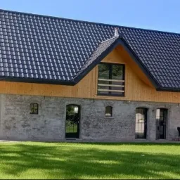 Nowoczesny dom z ciemną dachówką, drewnianą elewacją i szarym murem, otoczony zielonym trawnikiem.