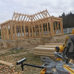 Ciesielstwo Polskie - Konstrukcja drewniana domu w trakcie budowy, widoczna piła do drewna DeWalt, przygotowane deski i porozrzucane kawałki drewna na ziemi.