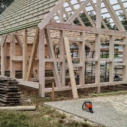 Konstrukcja szkieletowa domu w trakcie budowy, widoczne elementy drewniane, więźba dachowa i piła łańcuchowa na ziemi.