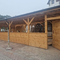 Ciesielstwo Polskie - Drewniana konstrukcja z zadaszeniem, widoczne elementy więźby, ściany z desek, lampa uliczna, kostka brukowa. Ujęcie z zewnątrz, w tle zieleń i budynek.