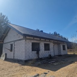Ciesielstwo Polskie - Budowa domu z bloczków: ściany wzniesione, dach pokryty ciemną dachówką, widoczna więźba dachowa. Na pierwszym planie piasek i narzędzia budowlane.