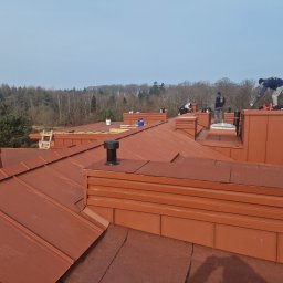 Ciesielstwo Polskie - Montaż brązowego dachu z blachy na budynku. Widoczni dekarze pracujący na dachu, kominy i las w tle. Ujęcie z góry, podkreślające strukturę i kolorystykę pokrycia.