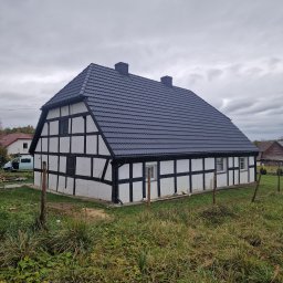 Ciesielstwo Polskie - Dom szkieletowy z ciemną dachówką w stylu rustykalnym, otoczony zielenią. Widoczne detale konstrukcyjne i kominy. Pochmurne niebo w tle.