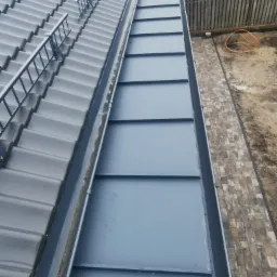 Szary dach z blachy trapezowej z zamontowanymi barierkami przeciwśniegowymi, widok z góry na obróbkę blacharską i chodnik z kostki brukowej przy ogrodzeniu.