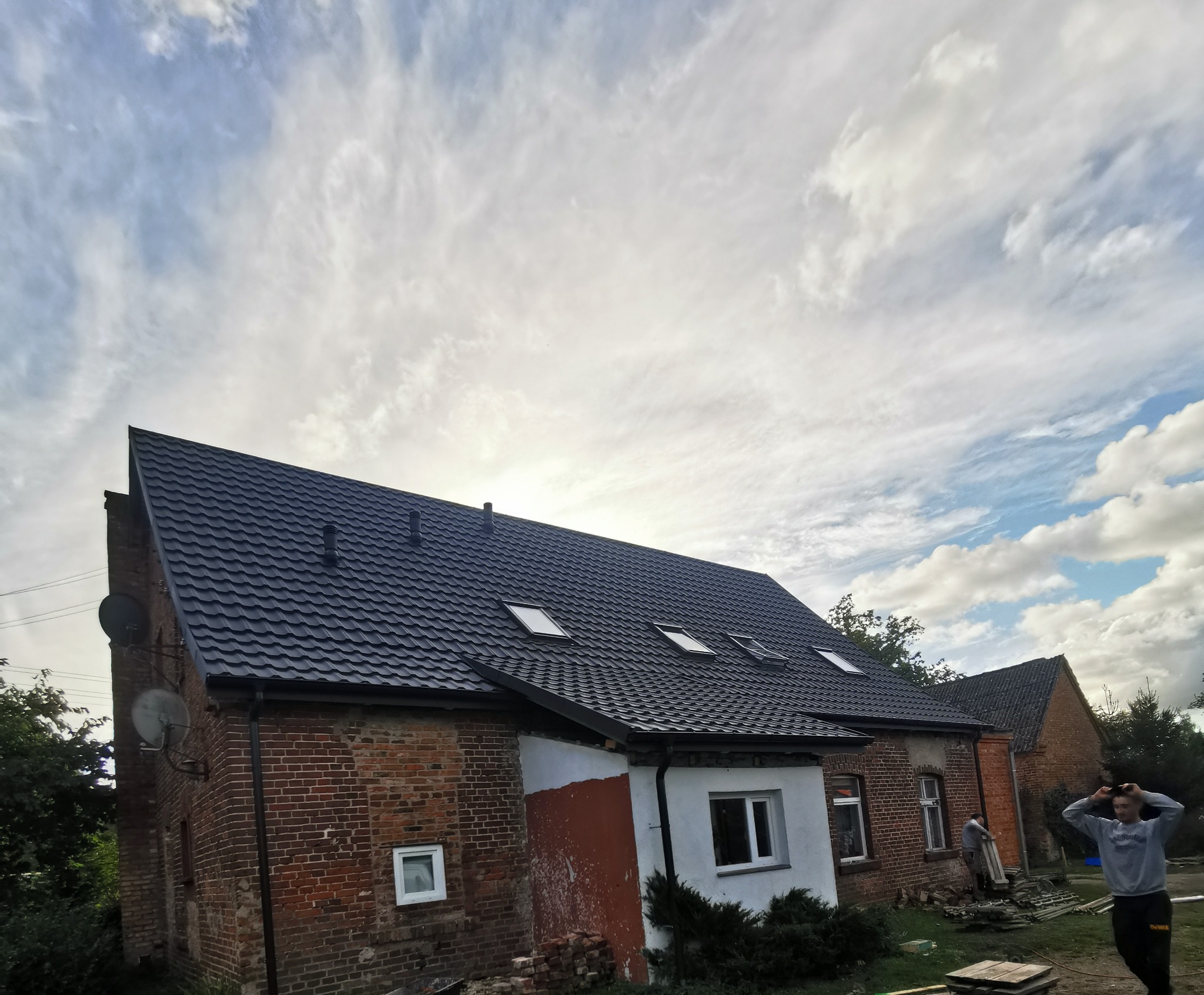 Odnowiony dach z dachówką bitumiczną na starym ceglanym budynku z oknami dachowymi, widoczne satelity i fragment nieotynkowanej ściany, na tle pochmurnego nieba.