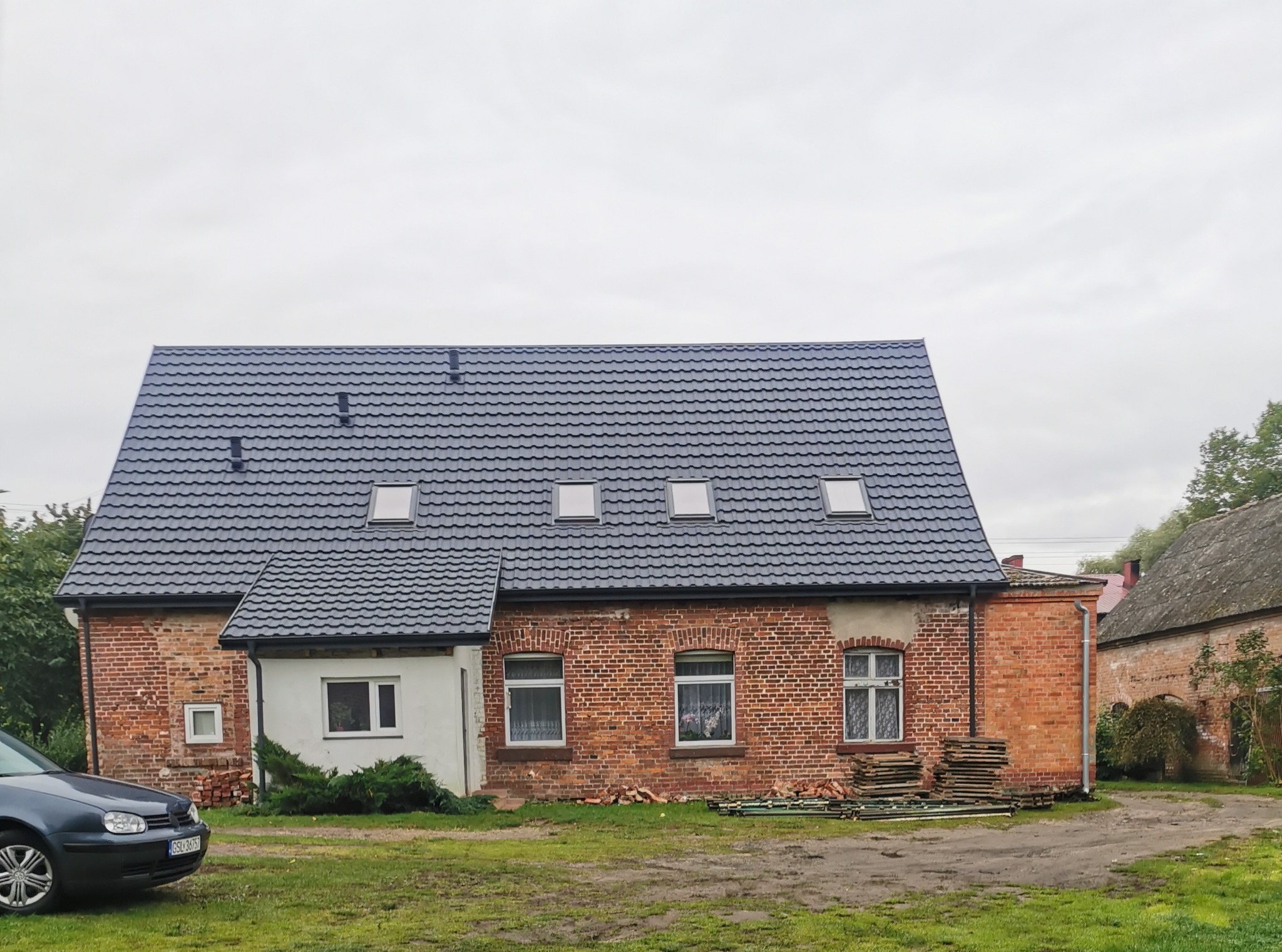 Ceglany dom z nowym, ciemnoszarym dachem z dachówki, widoczne okna dachowe, na podwórku stosy cegieł i zaparkowany samochód.