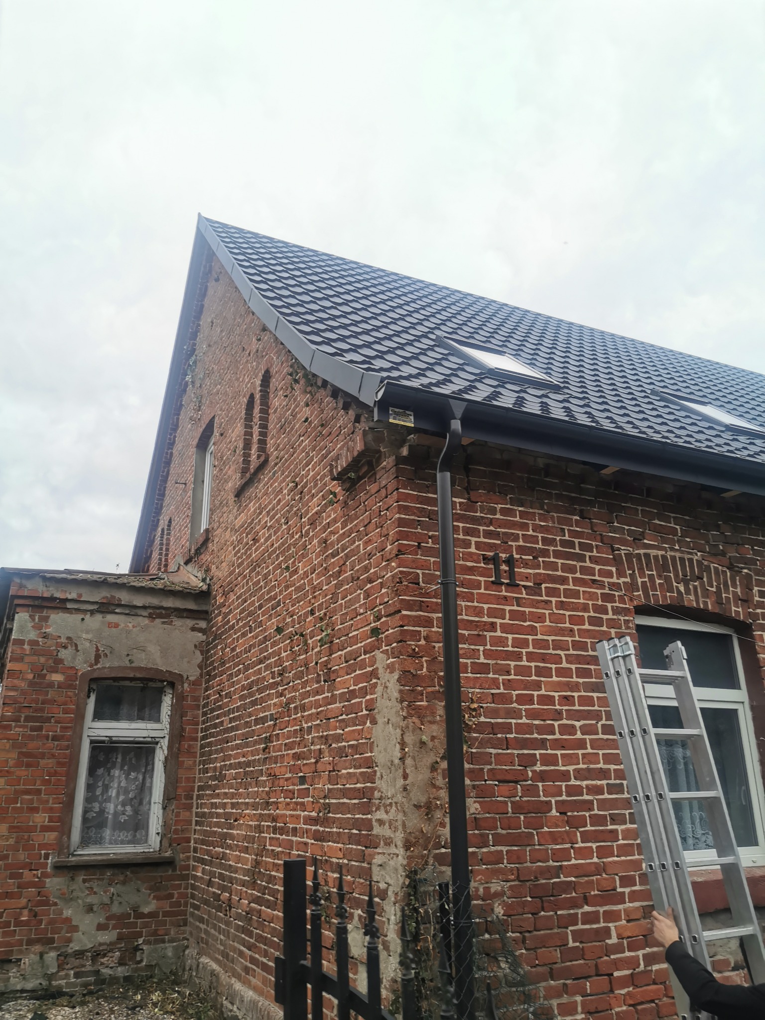 Ceglany budynek z nowym, ciemnym dachem z dwoma oknami dachowymi, widoczna drabina oparta o ścianę, zardzewiałe elementy elewacji i czarne numery '11' na ścianie.