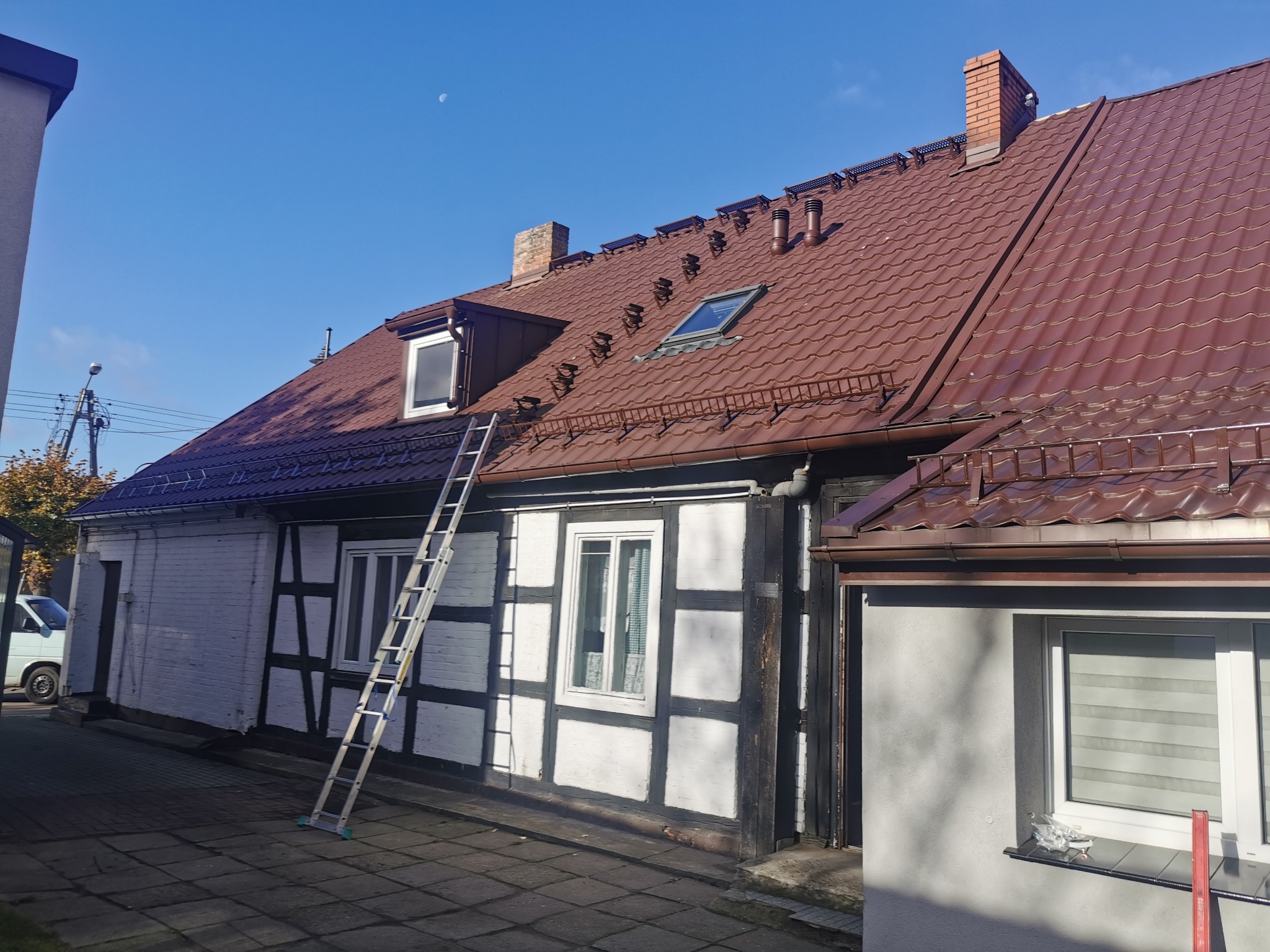 Dwukondygnacyjny budynek z muru pruskiego z nowym, brązowym dachem z dachówki, wyposażonym w okno dachowe i zabezpieczenia przeciwśniegowe, widziany w słoneczny dzień z drabiną opartą o ścianę.
