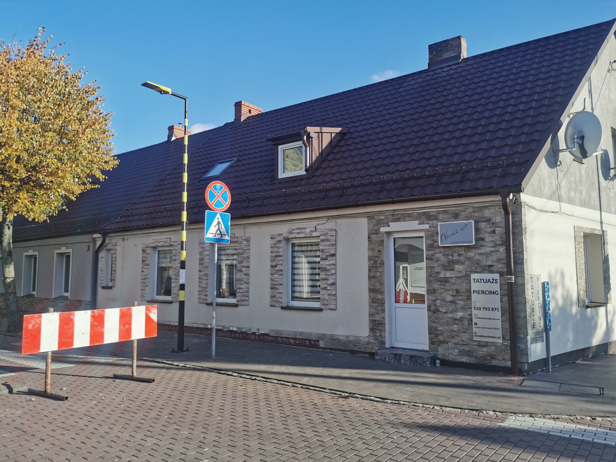 Dwukondygnacyjny budynek z ciemnobrązowym dachem, widoczne okna dachowe, komin i satelita. Przed budynkiem znak drogowy i bariera ostrzegawcza.