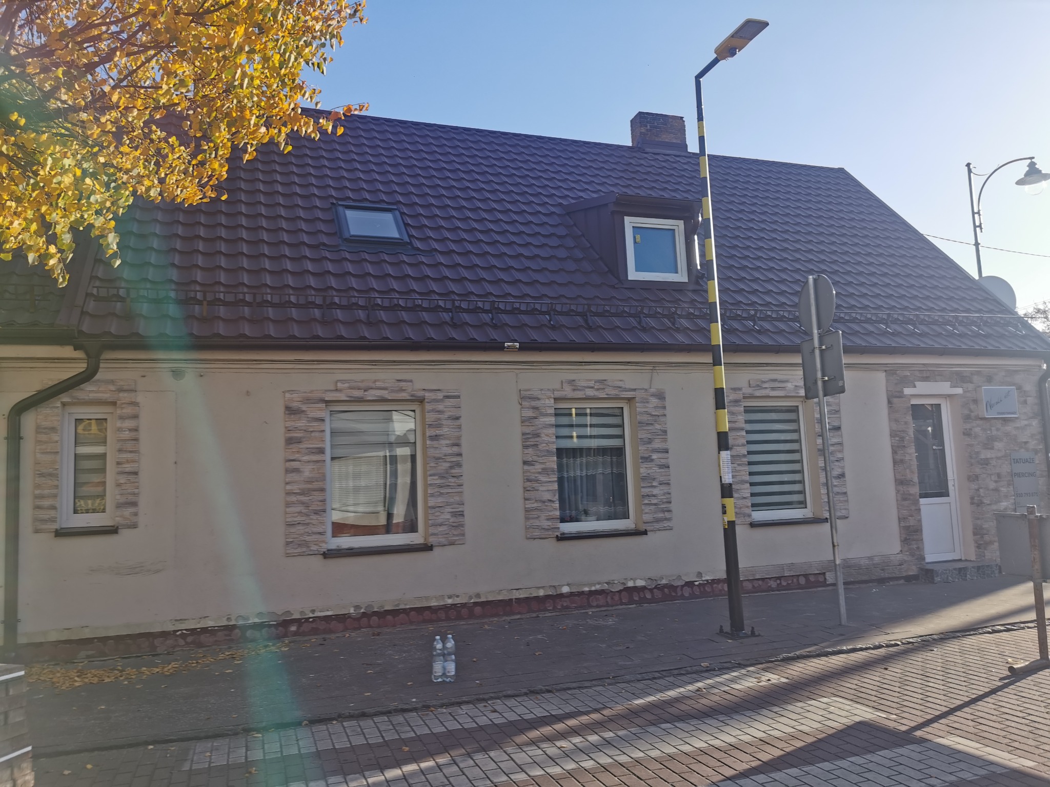 Dwukondygnacyjny budynek z ciemnobrązowym dachem z dachówką, oknami dachowymi i ceglaną elewacją, sfotografowany w słoneczny dzień z widocznymi drzewami z żółtymi liśćmi i butelkami wody na chodniku.