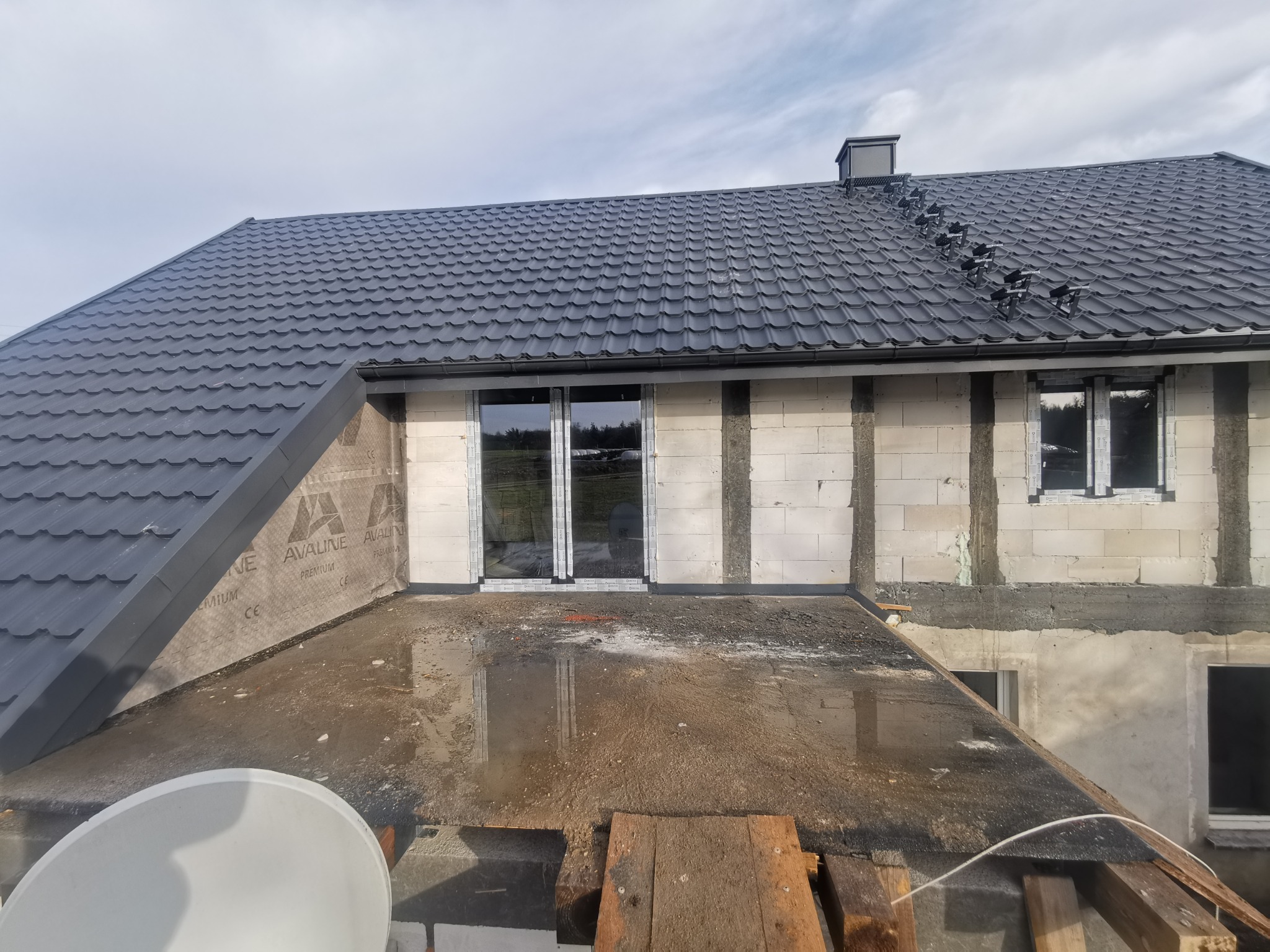 Nowy grafitowy dach z metalowej dachówki modułowej, zamontowane stopnie kominiarskie, ściany budynku w trakcie wznoszenia z betonu komórkowego, widoczne okna i folia wiatroizolacyjna.