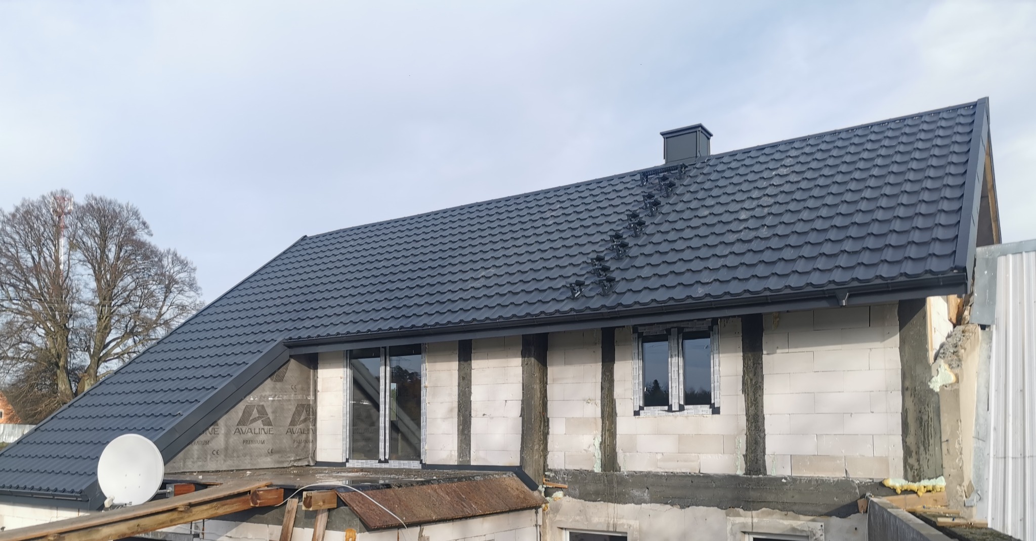 Nowy, grafitowy dach z blachy trapezowej na budynku w trakcie budowy, widoczne okna i surowy mur z betonu komórkowego, antena satelitarna na pierwszym planie.