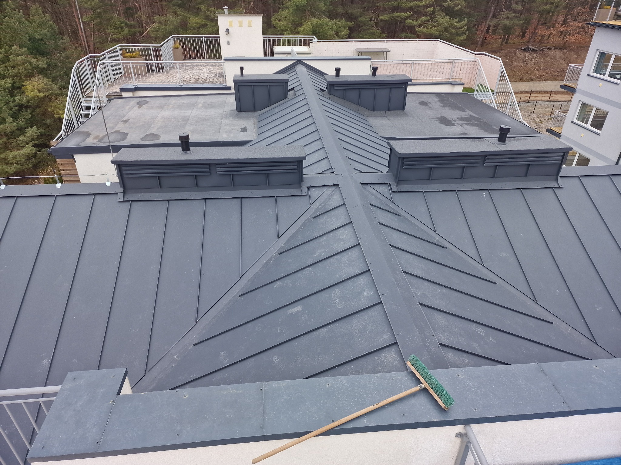 Widok z góry na szary, blaszany dach wielospadowy z lukarnami na nowoczesnym budynku wielorodzinnym; na pierwszym planie leży szczotka z długim, drewnianym trzonkiem.