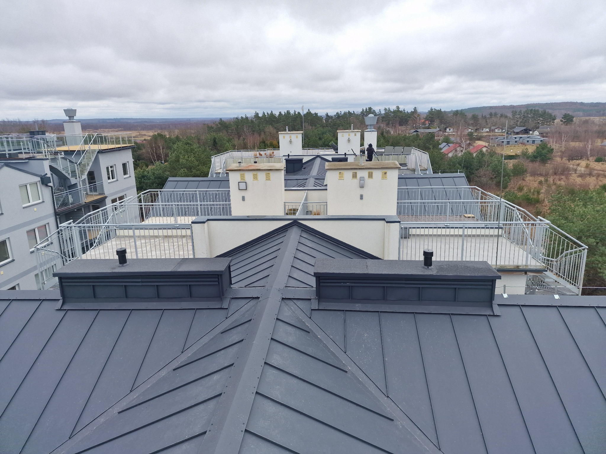 Szary dach z blachy na budynku wielorodzinnym, widok z góry na kominy i balustrady, w tle las i pochmurne niebo.