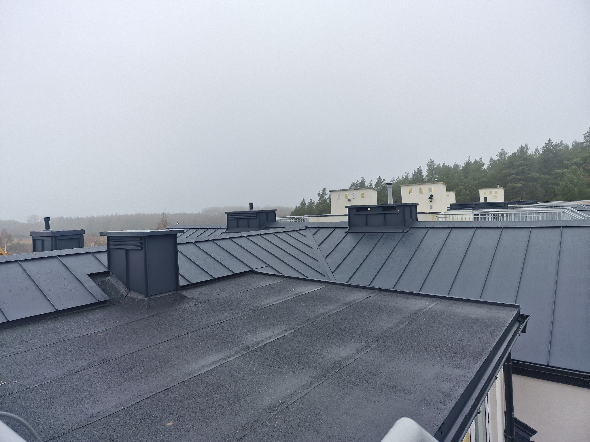 Czarny, płaski dach z blachy na budynku z kominami, widok z góry w pochmurny dzień.