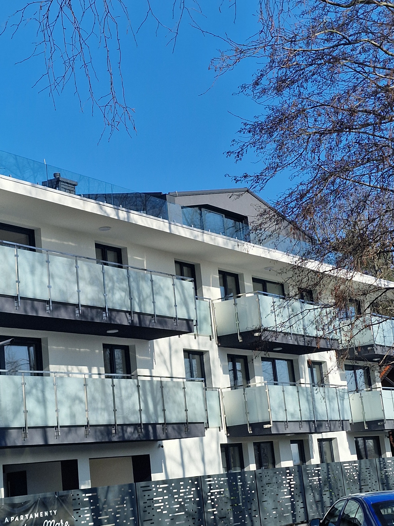 Nowoczesny budynek apartamentowy z białymi ścianami, czarnymi ramami okiennymi i balkonami z mlecznego szkła, widziany z dołu pod błękitnym niebem i gałęziami drzew