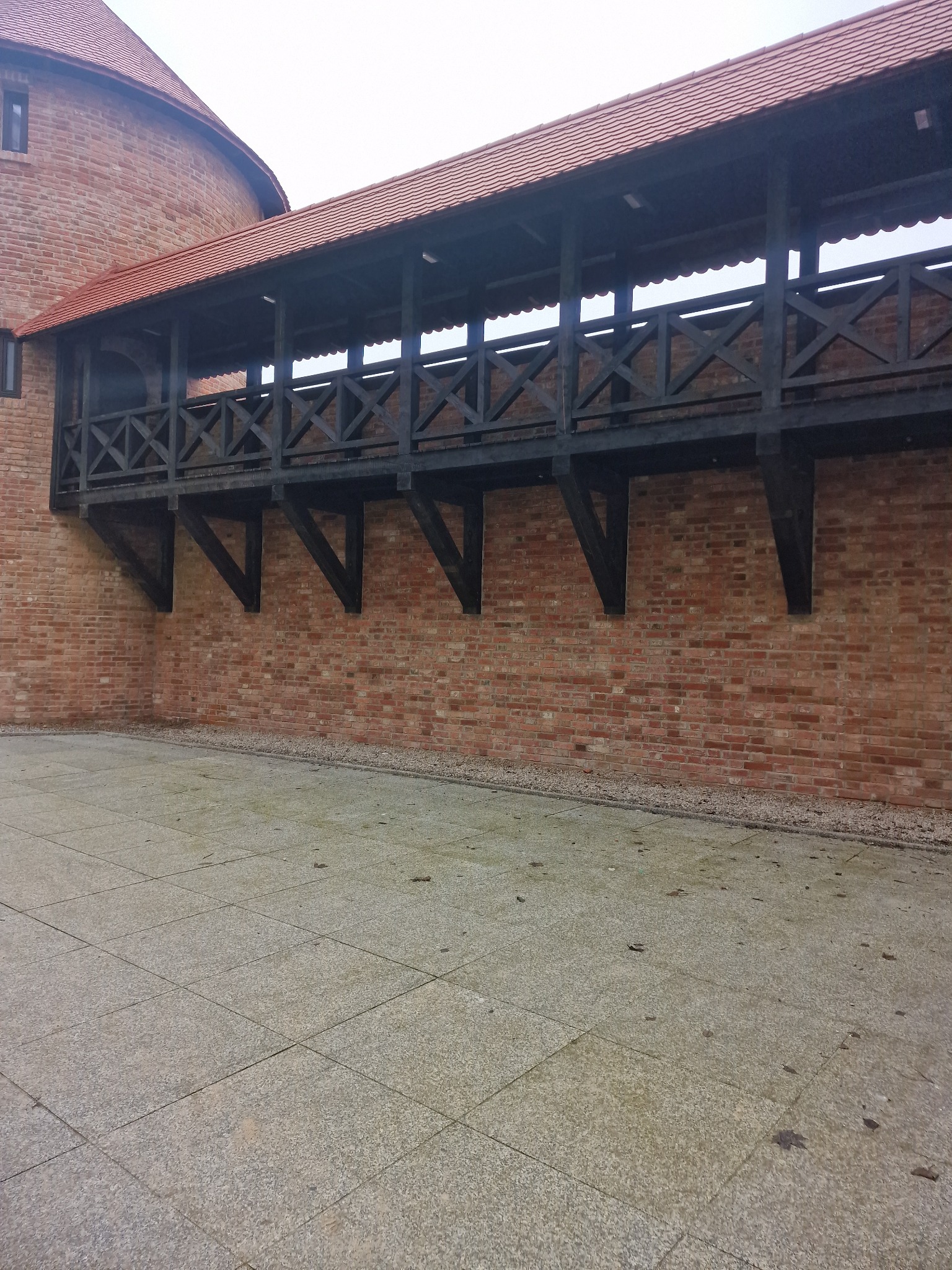 Ceglany budynek z drewnianym, zadaszonym przejściem wspartym na wspornikach, wykończonym czerwoną dachówką.